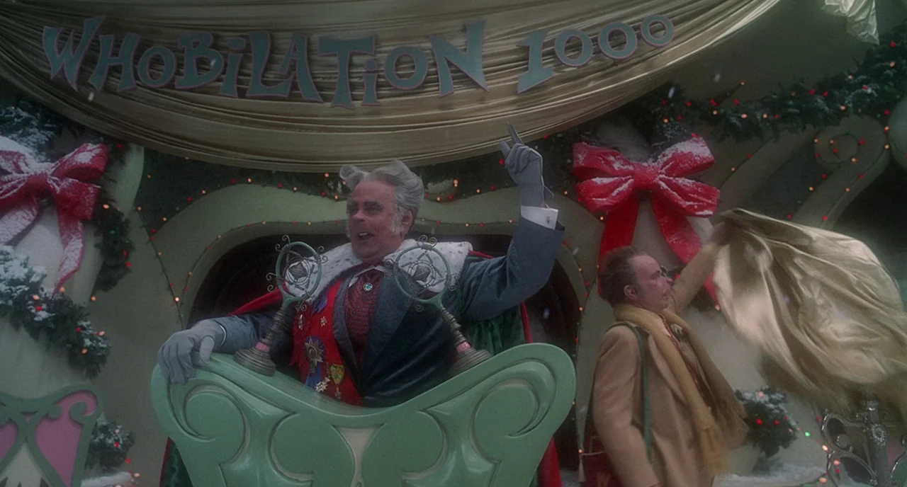 How The Grinch Stole Christmas (2000) movie still: medium — A whimsical, snow-dusted town square features a large banner displaying the text…