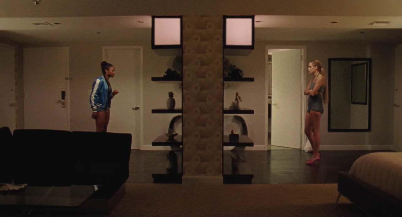 Zola (2020) movie still: fullbody — A modern hotel room with dark flooring features a central floor-to-ceiling wooden shelf divider. Two women stand…