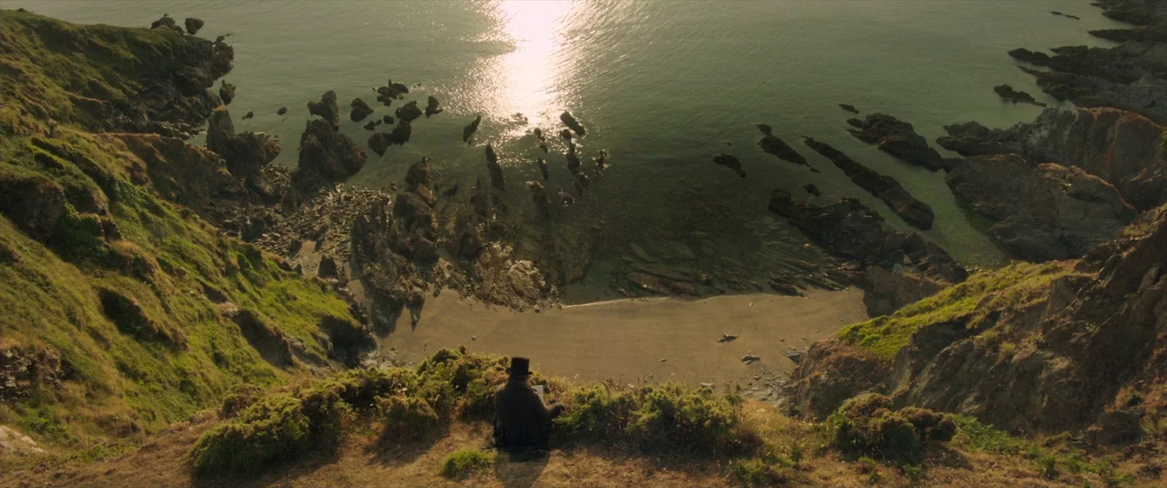 Mr. Turner (2014) movie still: wide — A grassy cliff overlooks a rocky beach and the sea under bright daylight. A person in a top hat sits on the…