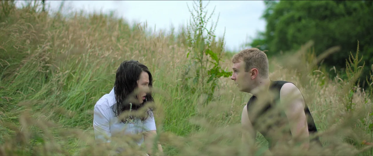 The Feast (2021) movie still: medium — A field of tall, unkempt green grass under a pale, overcast sky. A man and woman in simple clothing crouch in…