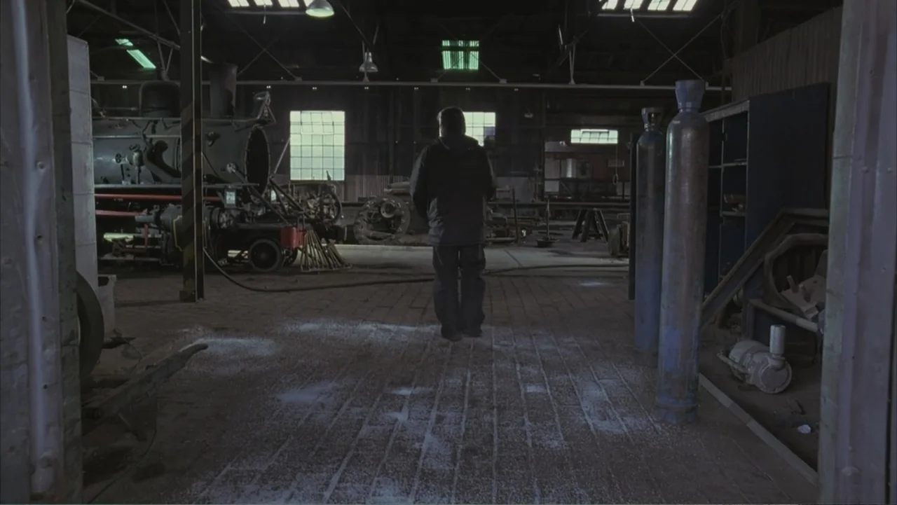 A Distant Place (2020) movie still: fullbody — A dark, industrial train shed contains a large steam locomotive and metal industrial machinery. A…