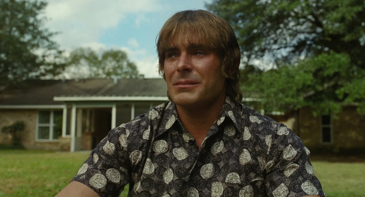 The Iron Claw (2023) movie still: medium — A suburban residential yard with a single-story brick home in the soft-focus background. A man with…
