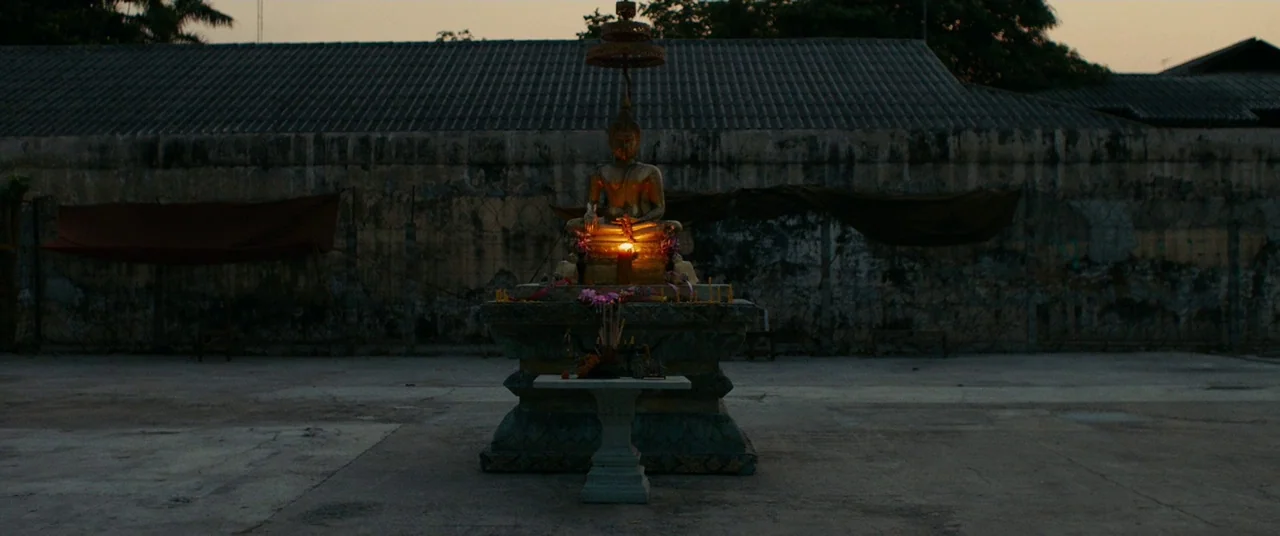 A Prayer Before Dawn (2017) movie still: wide — A stone Buddha statue sits on a tiered pedestal in a paved, open-air area. Dark fabric drapes hang…