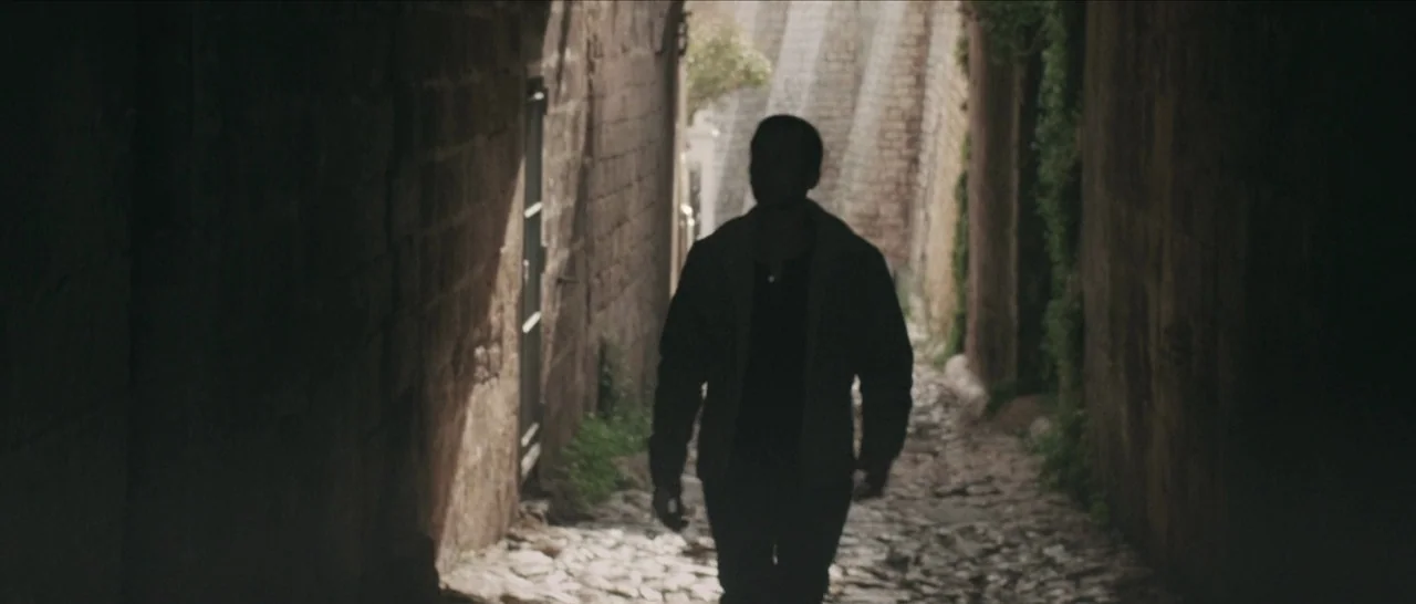 Spring (2014) movie still: fullbody — A narrow cobblestone walkway framed by high, aged stone walls in a shaded area. A man in dark clothing walks…