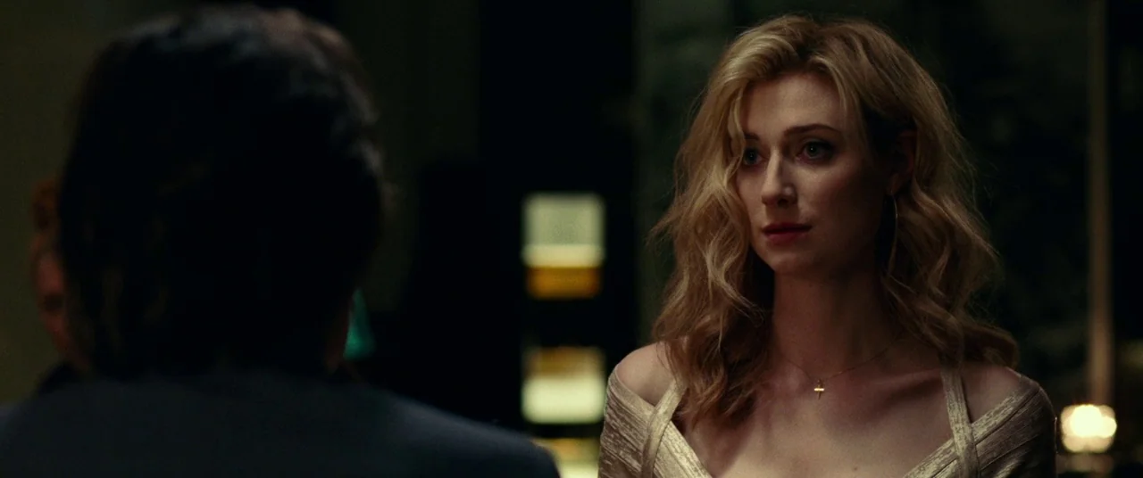 Widows (2018) movie still: medium — A dimly lit indoor space with blurred background elements featuring warm light sources. A blonde woman in a gold…