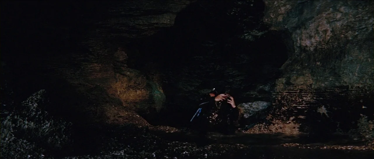 Thunderball (1965) movie still: wide — A dark, jagged cave interior features rough stone walls and a damp rocky floor. A scuba diver sits on the…