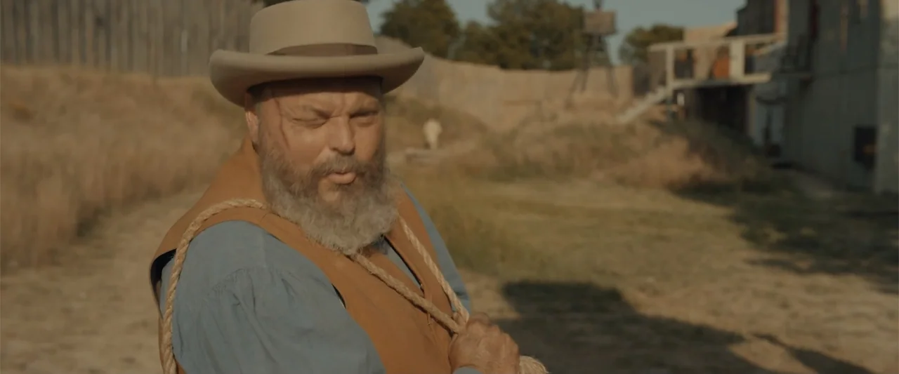 Ghosts of the Ozarks (2022) movie still: medium — A dusty, sunlit outdoor area features earthen fortifications and a wooden building structure. A…