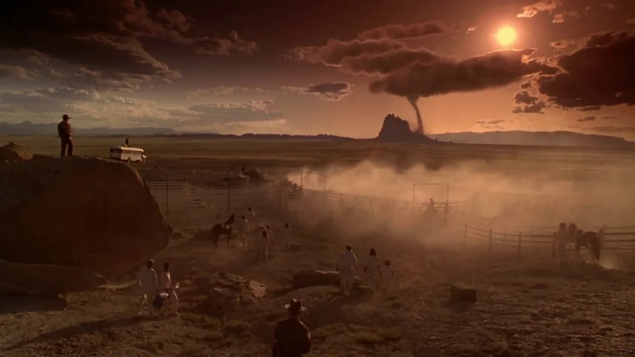Natural Born Killers (1994) movie still: wide — A vast, arid landscape features a distant mesa with a singular dark tornado cloud. A group of people…