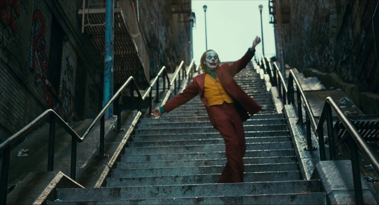 Joker (2019) movie still: fullbody — A long, steep concrete staircase in an urban alleyway is flanked by brick buildings. A man in a vivid red suit…