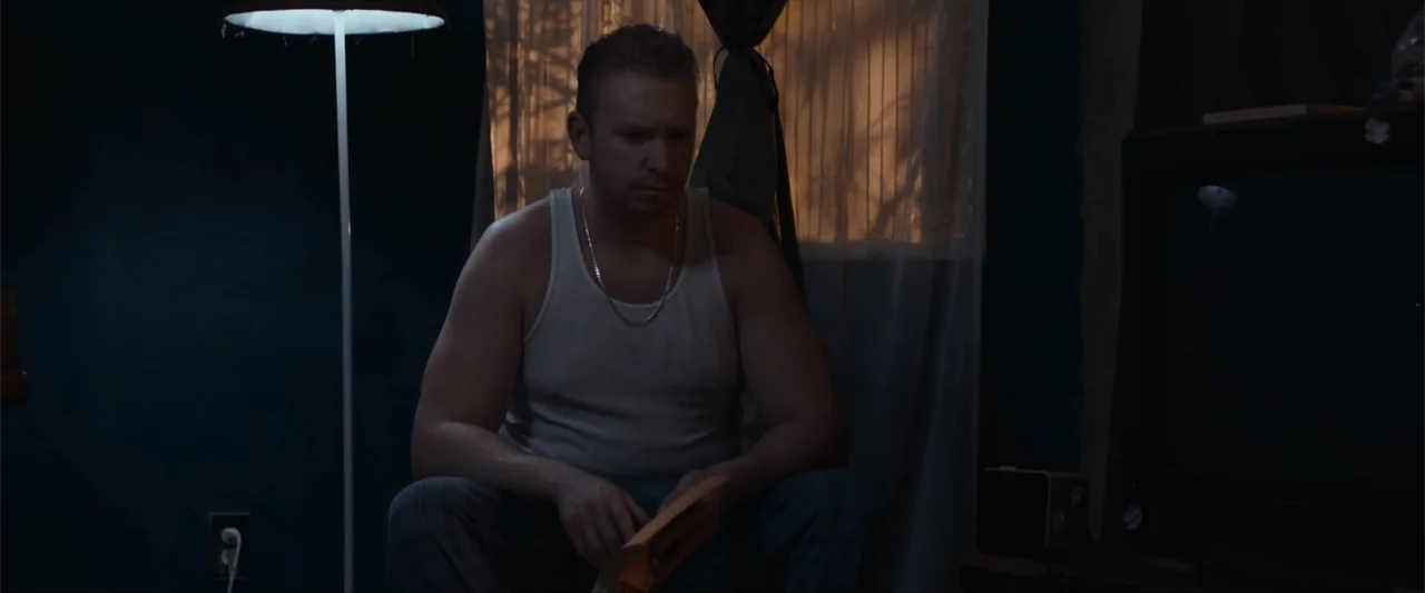 Anomaly (2006) movie still: medium — A dimly lit room features dark blue walls, a floor lamp, and a covered window. A man in a white tank top sits…