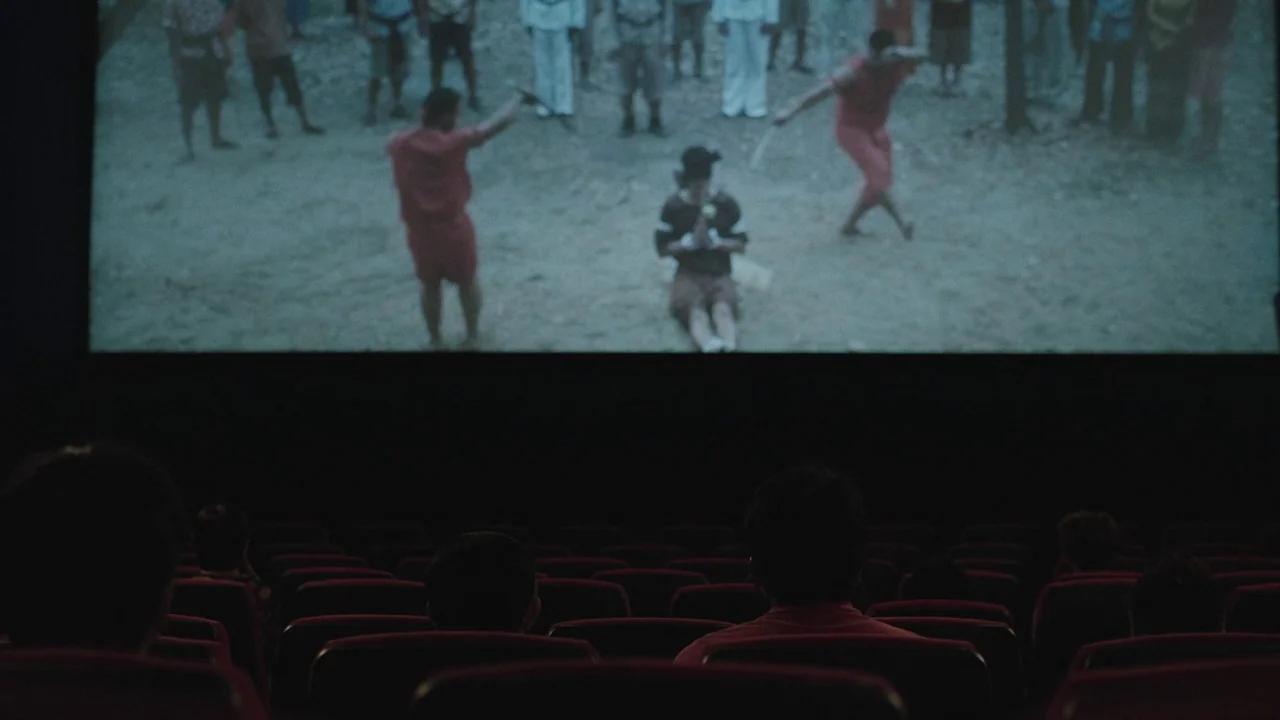 Cemetery of Splendour (2015) movie still: wide — A dark movie theater with rows of empty red seats facing a bright screen. Several audience members…