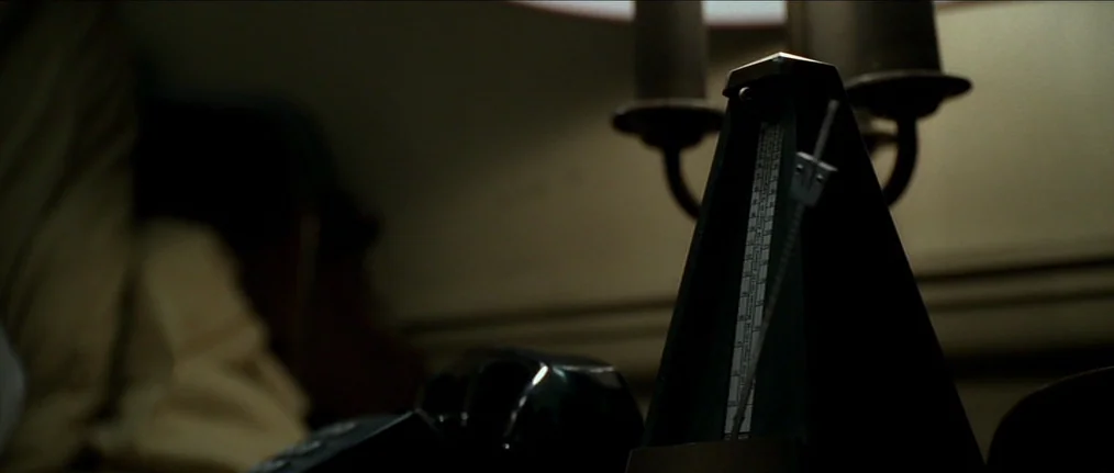 Se7en (1995) movie still: closeup — A dark, dimly lit indoor space contains a black metronome on a desk surface. The background features a blurry…