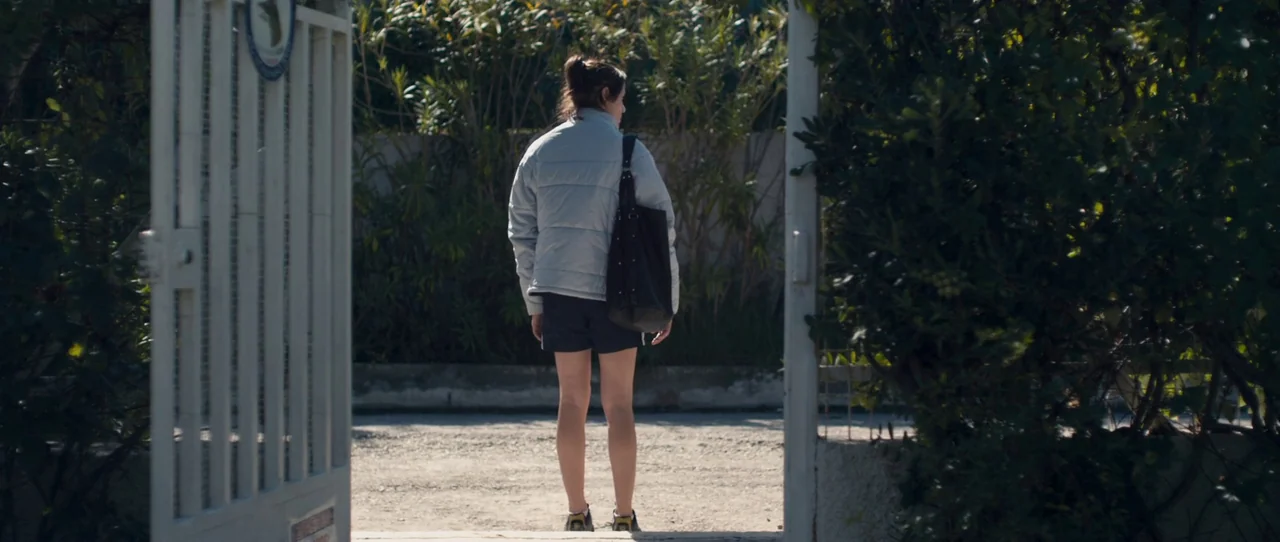 Alps (2011) movie still: fullbody — A person stands at a metal gate entrance in front of dense green bushes. The individual faces away from the…