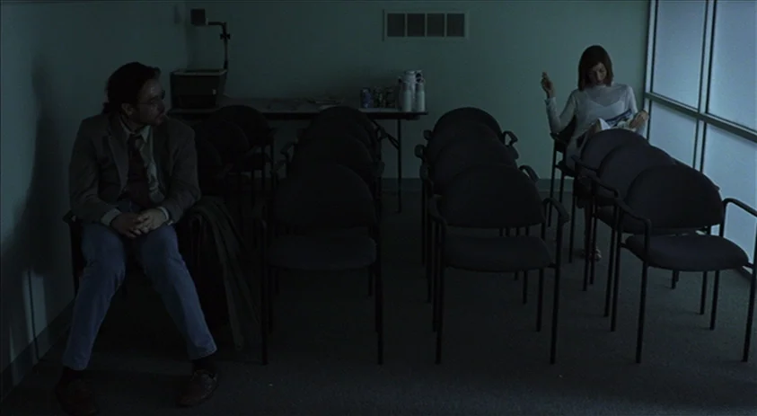 Being John Malkovich (1999) movie still: wide — A sparsely furnished waiting room has multiple rows of black chairs and a side table. A man sits on…