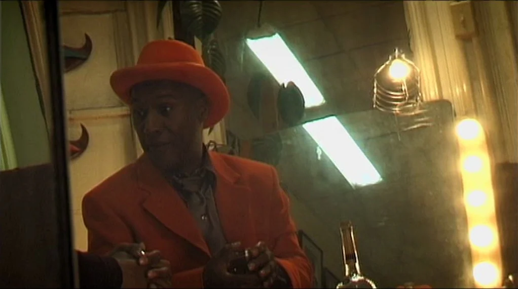 Bamboozled (2000) movie still: medium — A dimly lit room with mirrors and vanity lights reflecting the surroundings. A man in an orange suit looks…