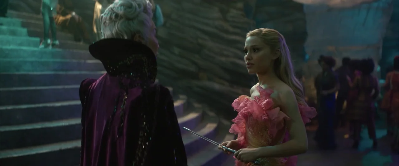 Wicked (2024) movie still: medium — A stone interior with tiered steps and textured walls serves as the backdrop. A woman in pink stands opposite a…