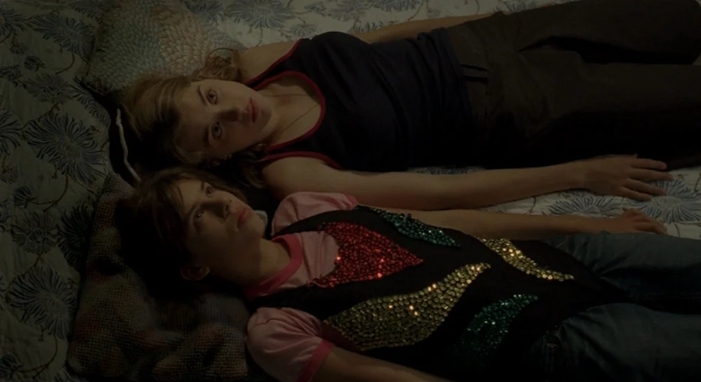 Water Lillies (2007) movie still: medium — A top-down view shows two women lying on a bed with a floral patterned comforter. They are positioned…
