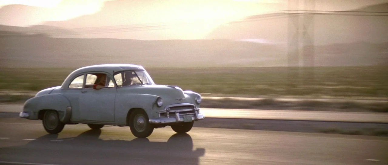 Starman (1984) movie still: wide — A vintage 1950s sedan drives along an open highway through a vast desert. Mountains rise in the soft-lit distance…