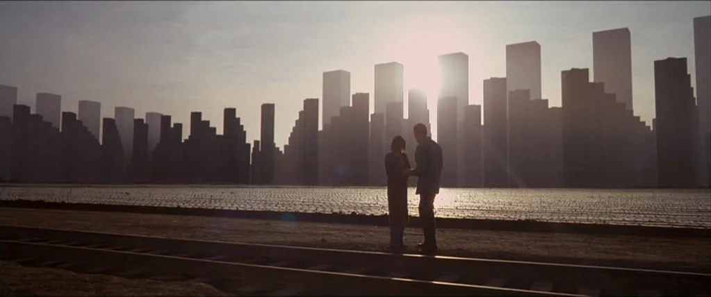 Inception (2010) movie still: wide — A man and woman stand on railroad tracks before an immense, hazy, stylized urban skyline. The sun sets directly…