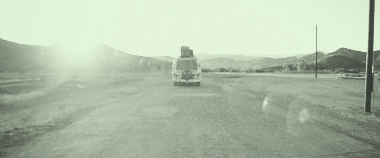 31 (2016) movie still: wide — A van travels down a desolate paved road through an open, flat desert area. Hazy mountains sit on the horizon under a…