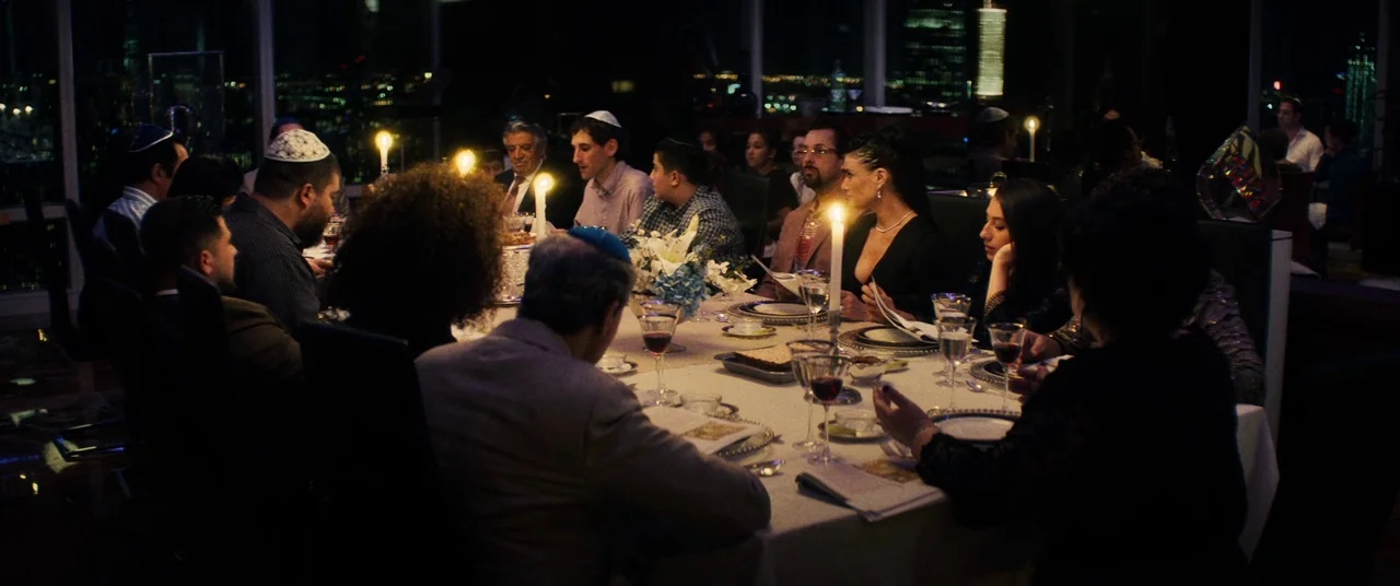 Uncut Gems (2019) movie still: wide — A group gathers at a long dinner table inside a room overlooking city lights. They participate in a formal…