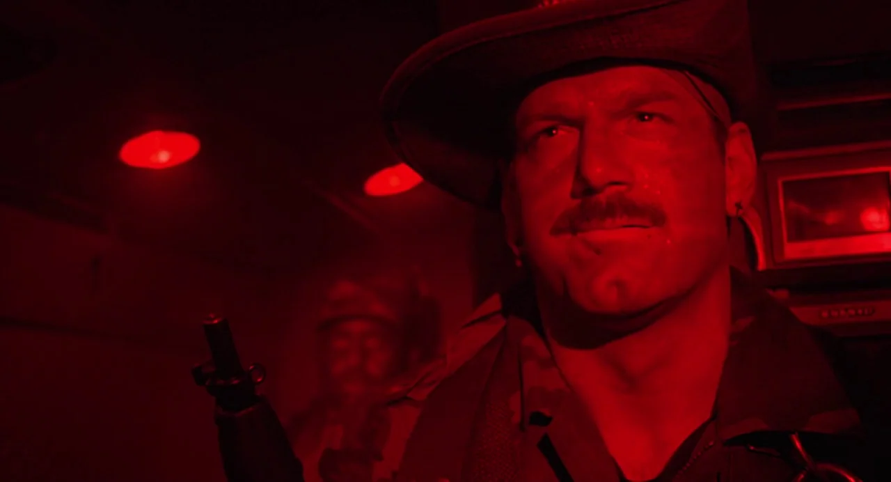 Predator (1987) movie still: medium — A dark, cramped military transport interior is bathed in deep red emergency light. A soldier with a mustache…