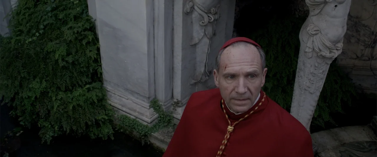 Conclave (2024) movie still: medium — A stone courtyard features classical marble carvings and lush green ferns in the background. An elderly man in…