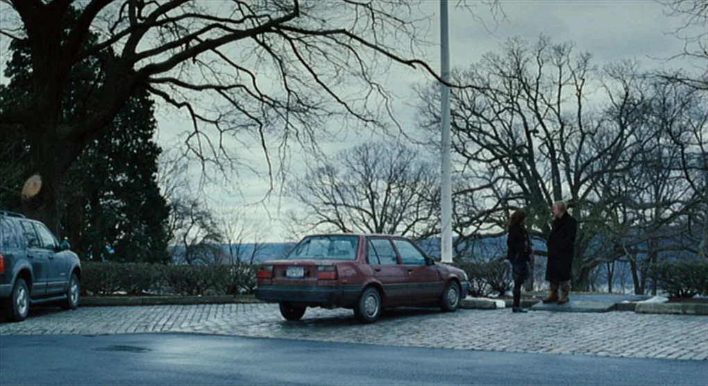 The Savages (2007) movie still: wide — A stone-paved parking lot borders bare trees and a gray sky on a winter day. A man and woman stand beside a…