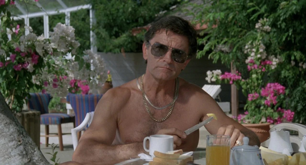 American Ninja (1985) movie still: medium — An outdoor patio area features blooming bougainvillea plants and white patio furniture. A shirtless man…