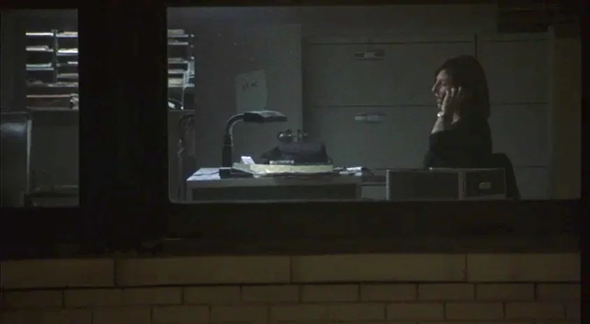 Being John Malkovich (1999) movie still: medium — A dark office environment viewed through an exterior window at night. A woman sits at a desk…
