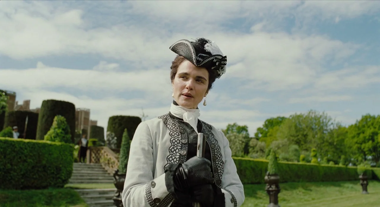 The Favourite (2018) movie still: medium — A sprawling formal garden with manicured hedges and stone steps leads toward a manor. A woman dressed in…