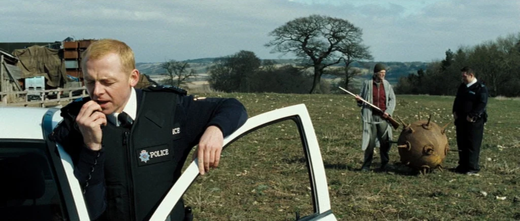 Hot Fuzz (2007) movie still: medium — An open grassy meadow with a bare tree stands under an overcast sky. A police officer radios in next to his…