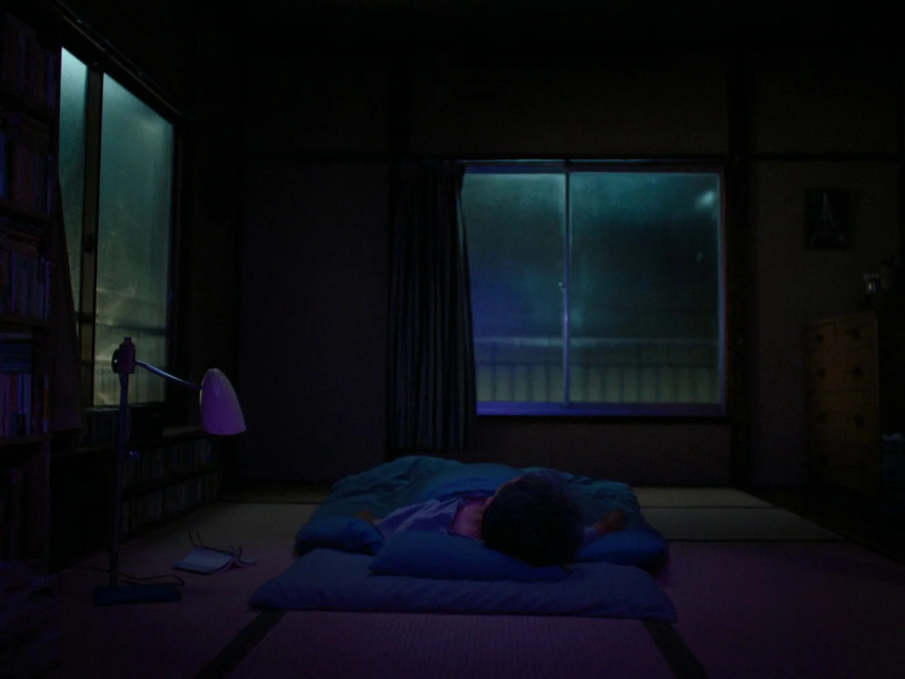 Perfect Days (2023) movie still: wide — A Japanese room at night with a single person sleeping on a floor futon. Low blue moonlight from the windows…