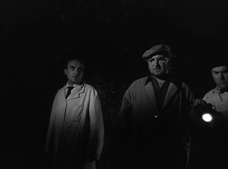 Head Against The Wall (1959) movie still: medium — A dark outdoor space at night with dense foliage visible behind three men. The men stand…