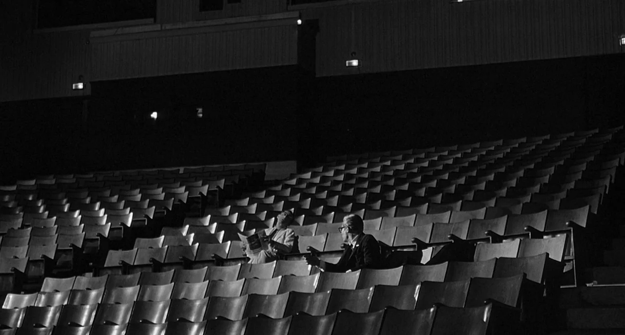 8 1/2 (1963) movie still: wide — A vast, dark cinema auditorium features rows of empty seats under dim ambient light. Two men sit in the middle of…