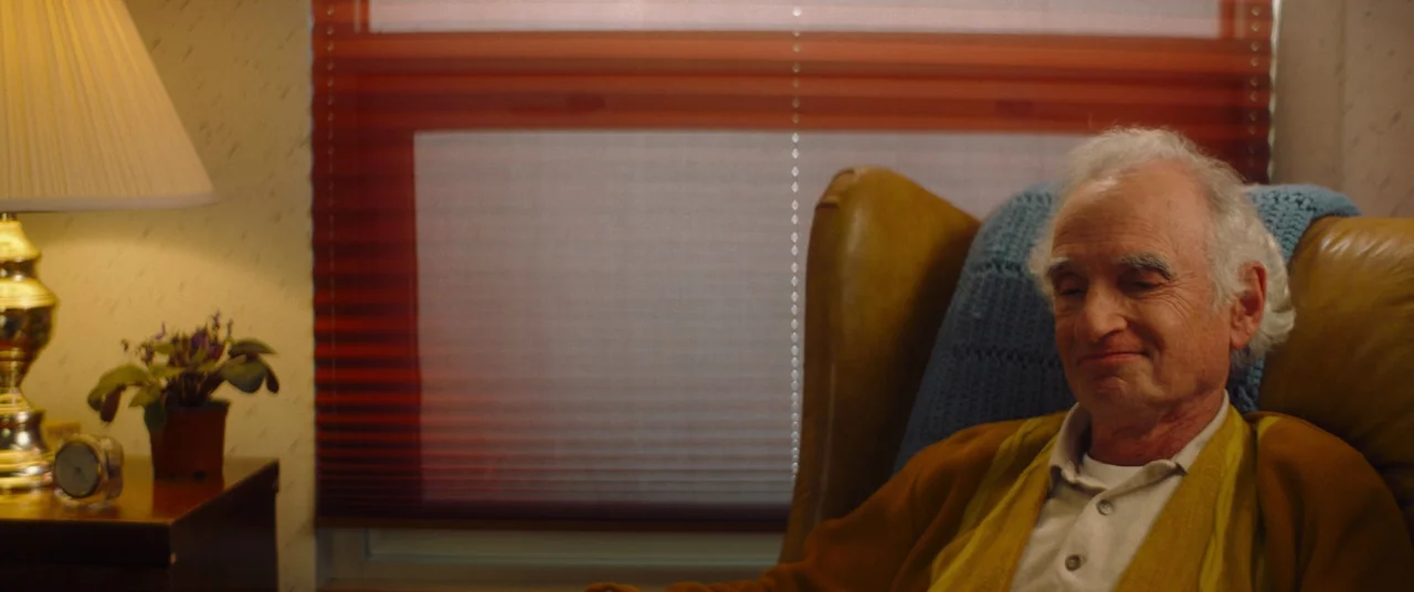 Linoleum (2022) movie still: medium — A domestic space features a window with orange pleated blinds and a side table. An elderly man sits in a brown…