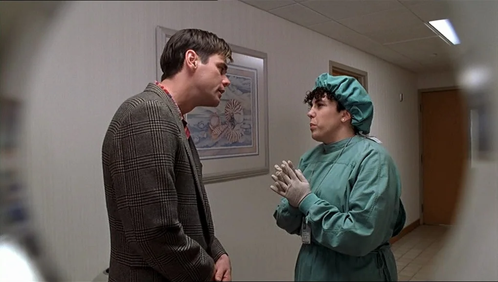 The Truman Show movie still: medium — A sterile hallway featuring beige walls and framed artwork depicting seashells. A man wearing a plaid blazer…