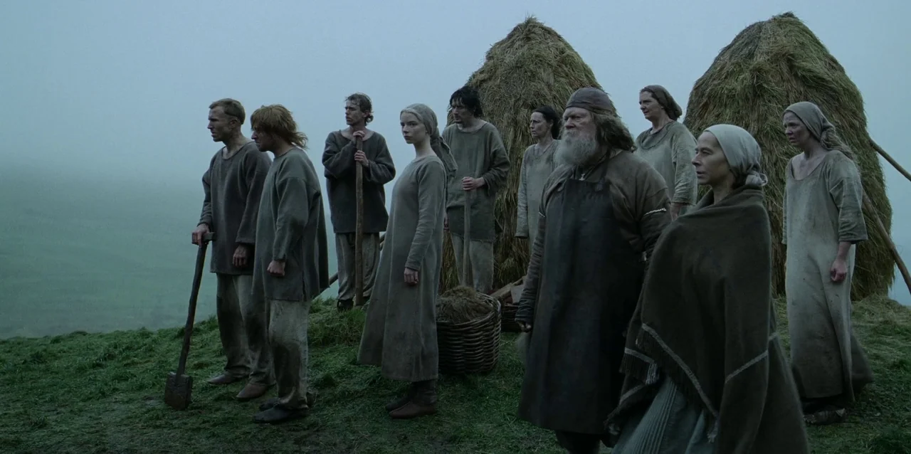 The Northman (2022) movie still: wide — A group of medieval peasants stands on a damp, green hillside near haystacks. They look out across a…