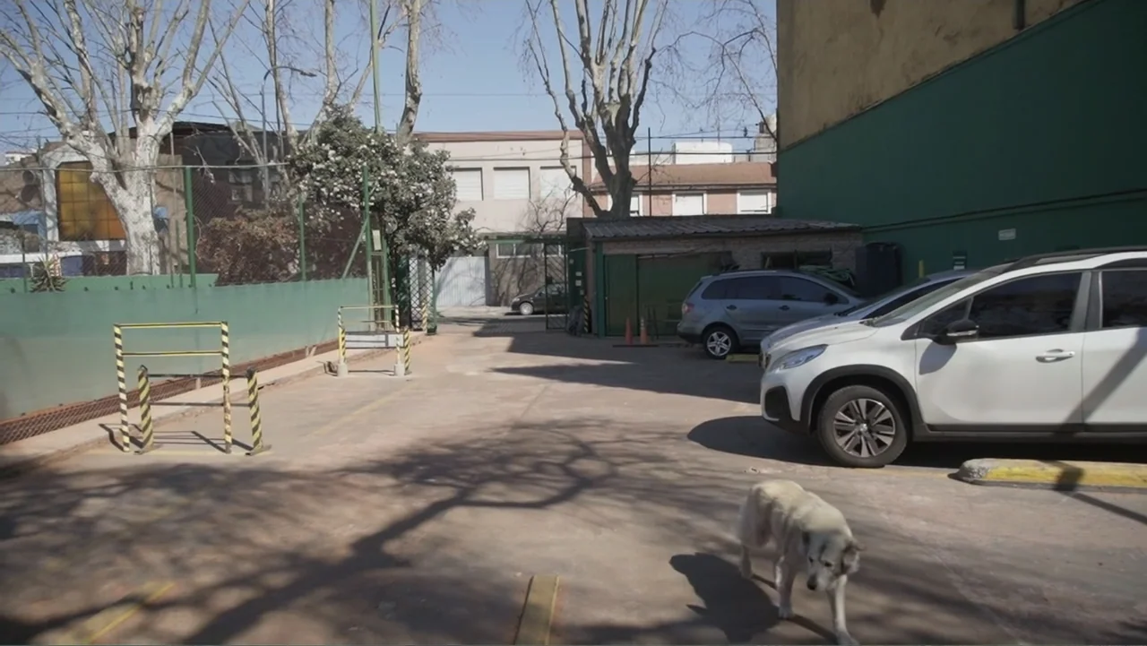 Saturdays Disorders (2022) movie still: wide — An outdoor concrete parking area bordered by a green metal fence and parked vehicles. A light-colored…