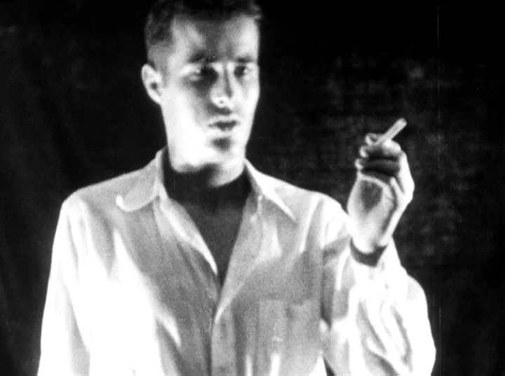 Fireworks (1947) movie still: medium — A dark space with a solid black wall background. A man stands wearing a white shirt while holding a cigarette…