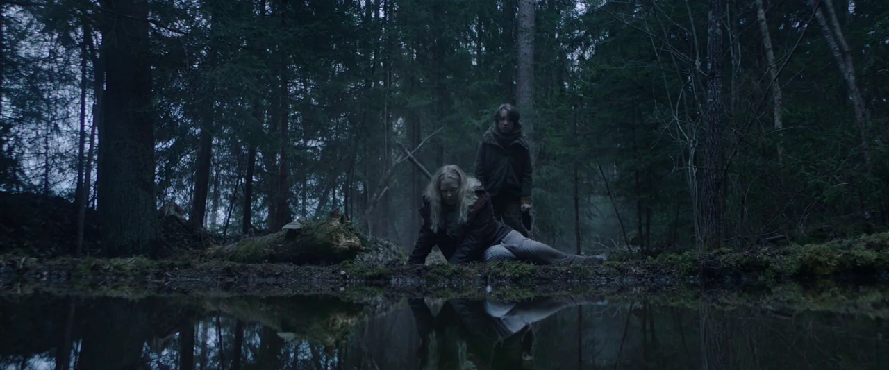 Vesper (2022) movie still: wide — A dense, overgrown coniferous forest surrounds a stagnant dark pool of water. Two figures are positioned near the…