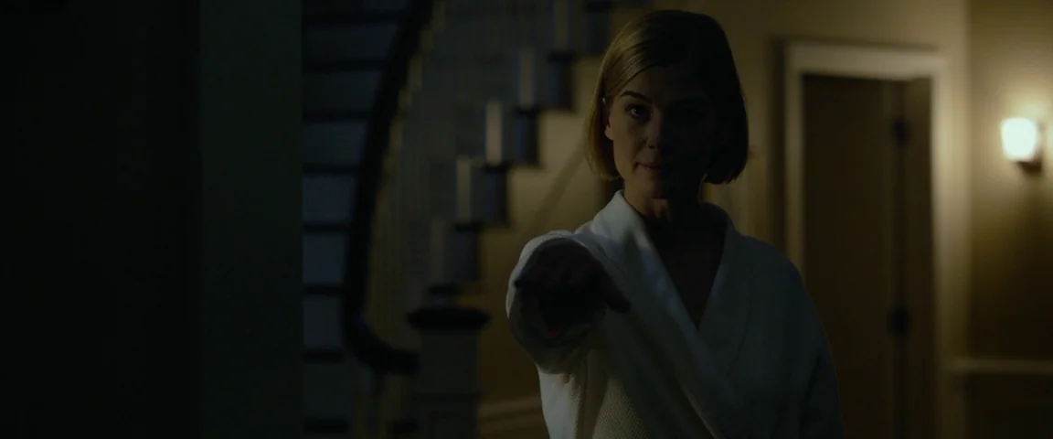 Gone Girl (2014) movie still: medium — A dark staircase landing in a residential home features a wooden banister. A woman stands in the shadows…