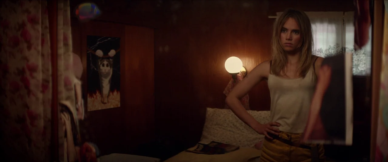 The Bad Batch (2016) movie still: medium — A dim room features wood paneling on the walls and a cat-themed poster pinned up. A young woman stands…