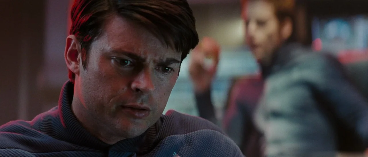 Star Trek (2009) movie still: closeup — A dimly lit interior space with futuristic dark technical panels and glowing red accents. A man in a dark…