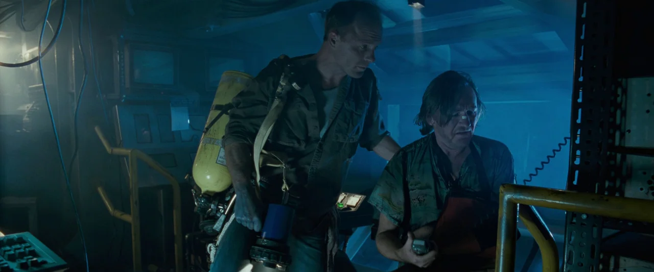 The Abyss (1989) movie still: medium — A dark, cramped industrial space filled with technical equipment and blue ambient light. Two men stand in the…