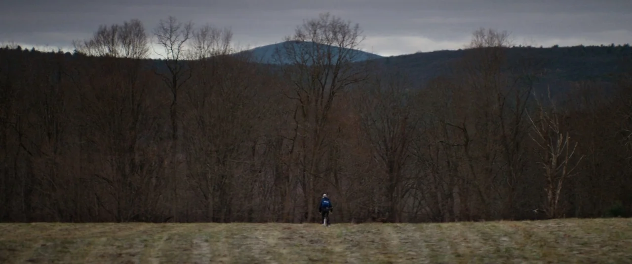 Super Dark Times (2017) movie still: wide — A vast, harvested field extends toward a dense line of leafless trees and mountains. A lone cyclist…