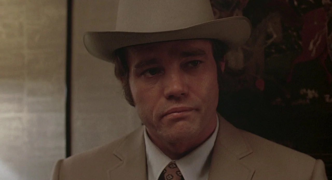 Charley Varrick (1973) movie still: closeup — An indoor space features a wall with a dark framed art piece. A man wearing a fedora and suit stands…