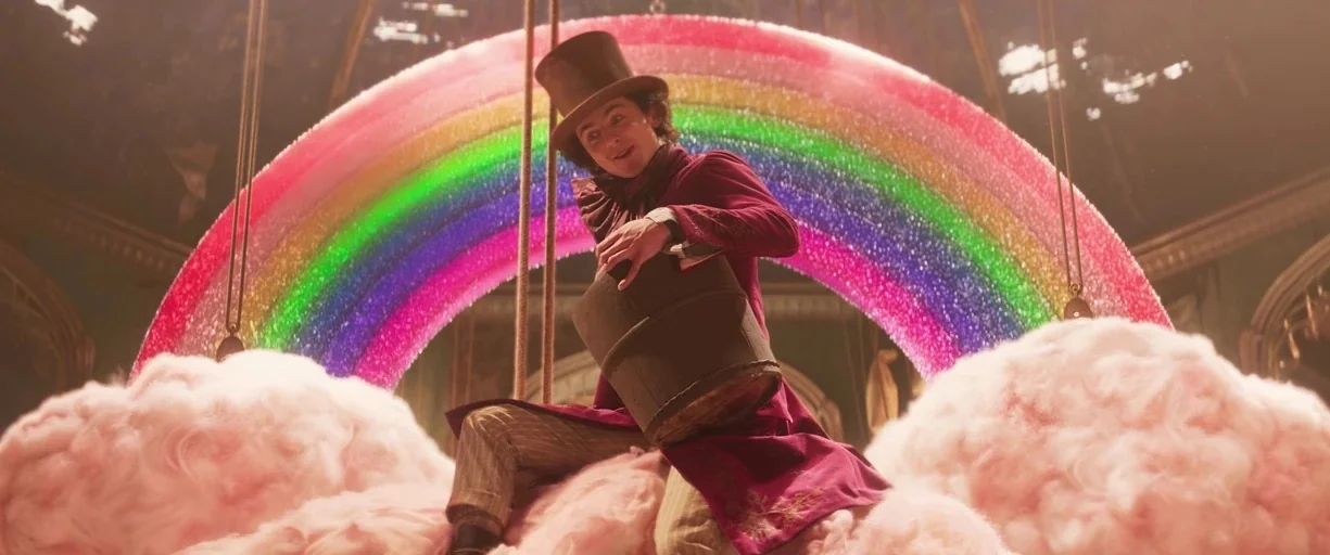 Wonka (2023) movie still: medium — A whimsical fantasy interior features a large glowing rainbow behind a person on clouds. The character sits atop…
