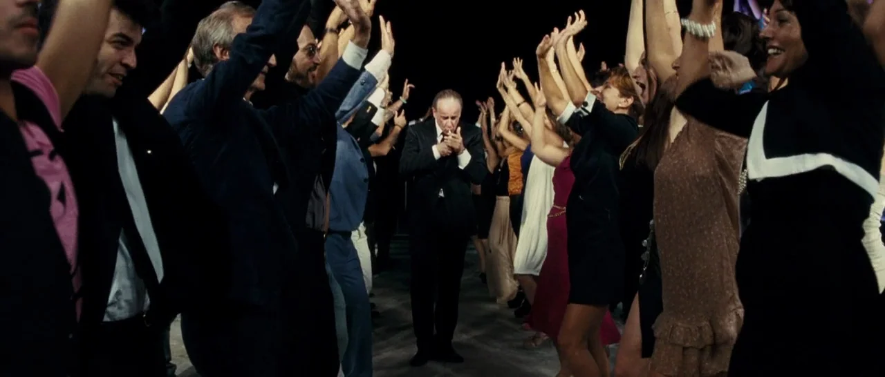 The Great Beauty (2013) movie still: fullbody — A dark, expansive indoor space features a crowd of people dancing with raised arms. A single man in…