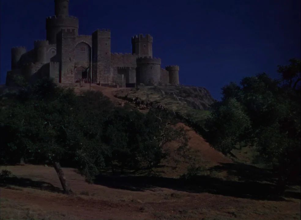 The Adventures of Robin Hood (1938) movie still: establishing — A stone castle sits atop a dark hill under a clear night sky. A dirt path winds up…